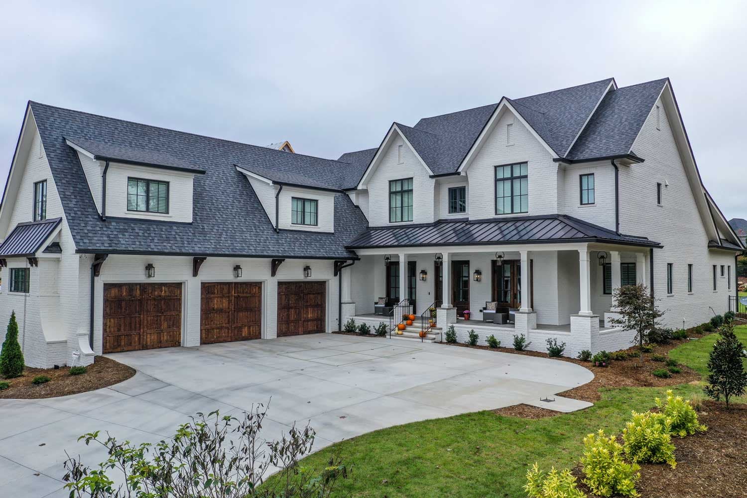 A large custom white-brick home with a dark shingle roof, a standing-seam metal porch roof, three carriage-style wood garage doors, steep gables, and a covered front porch