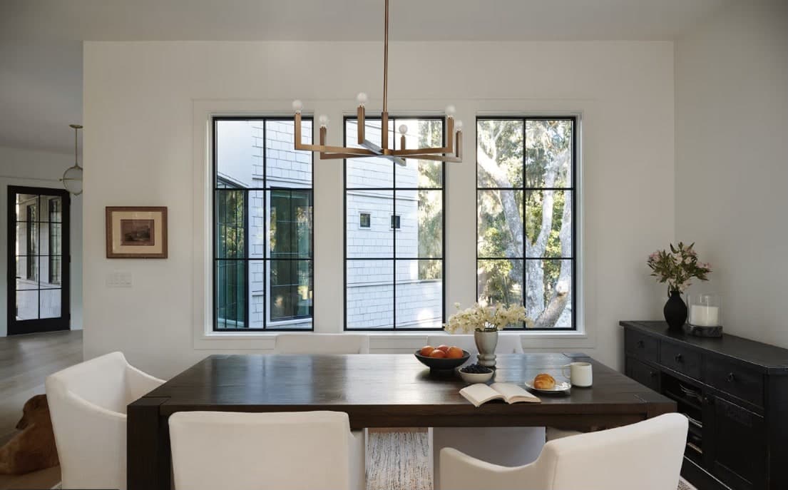Three black-framed divided-light windows in a modern farmhouse dining room, looking out to a shingle-sided coastal home and live oaks