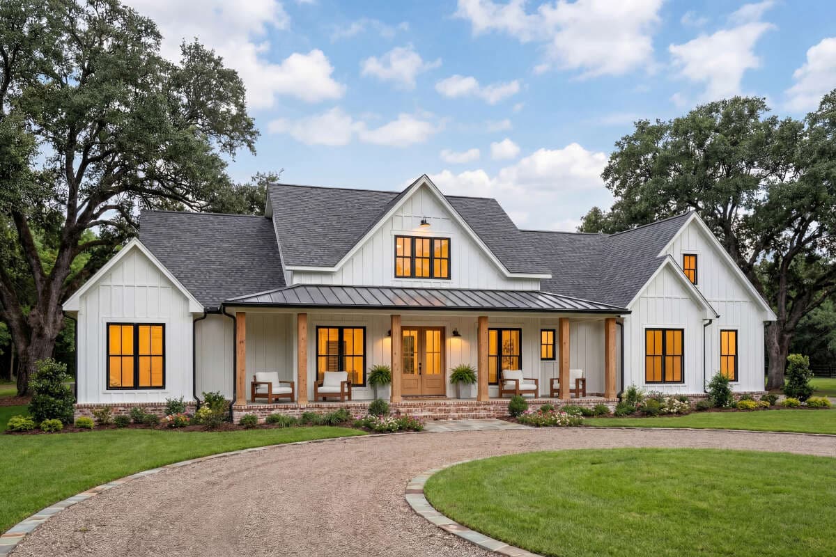 A white modern farmhouse with black-framed windows, board-and-batten siding, a standing-seam metal porch roof, and cedar porch posts, set behind a circular gravel drive and framed by mature oaks